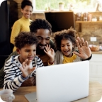 Father and children wave at a laptop webcam, mother smiles at them in the background.